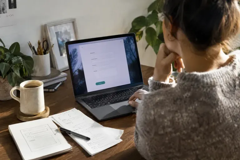 Designer reviewing a simple contact form prototype on a laptop, paper sketches of field labels nearby.